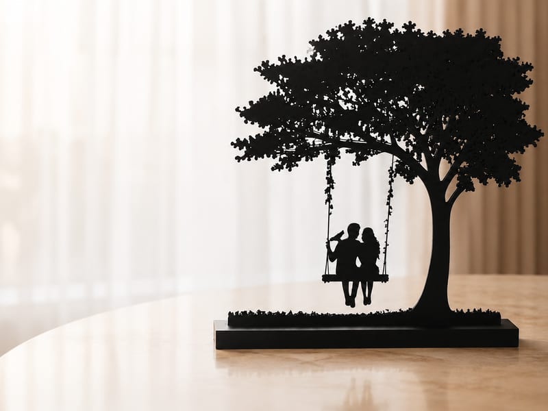 A black 3D-printed silhouette of a couple sitting on a swing beneath a full leafy tree, mounted on a Le Layers branded base, standing on a marble table.