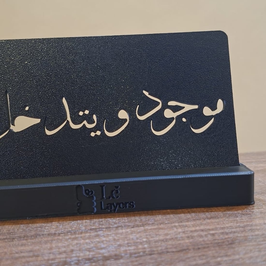 A matte black 3D-printed standing plaque with the Arabic phrase موجود و يتدخل cut through it and backed in gold, on a Le Layers branded base.