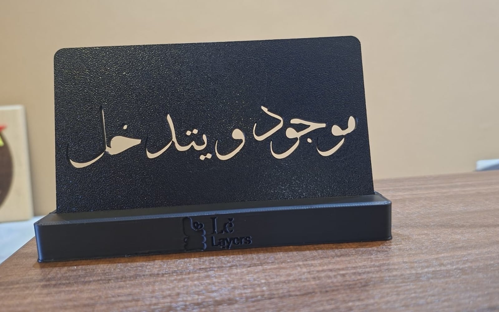 A matte black 3D-printed standing plaque with the Arabic phrase موجود و يتدخل cut through it and backed in gold, on a Le Layers branded base.