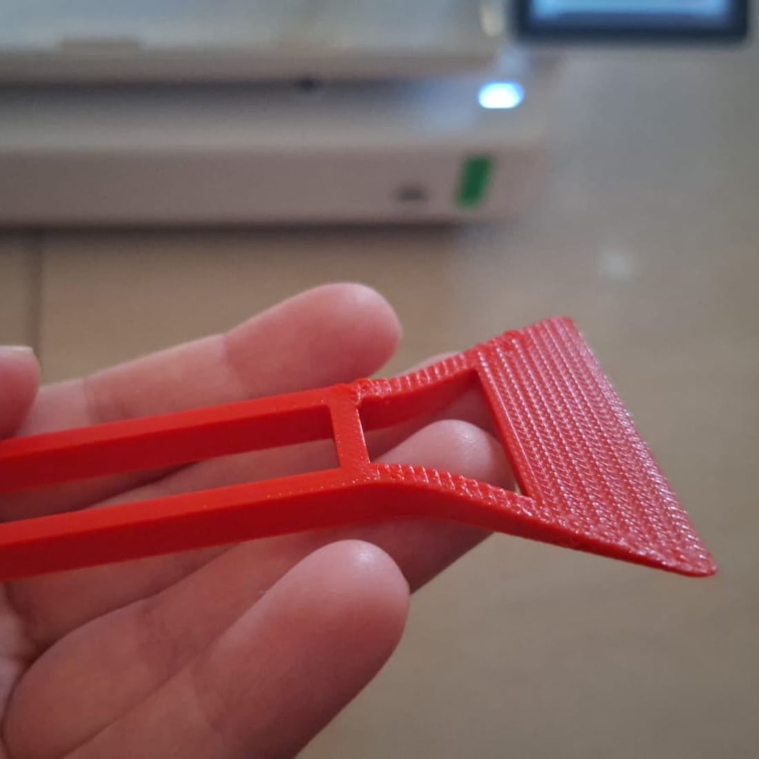 A red 3D-printed bed scraper tool held in a hand in front of a 3D printer.