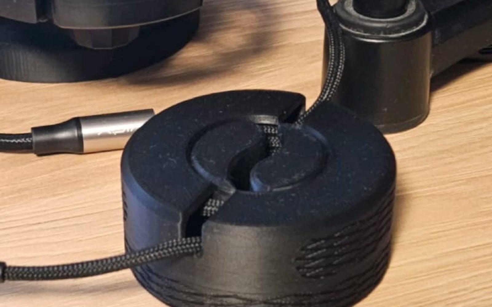 A black 3D-printed circular cable winder on a wooden desk with a braided microphone cable wound into it.