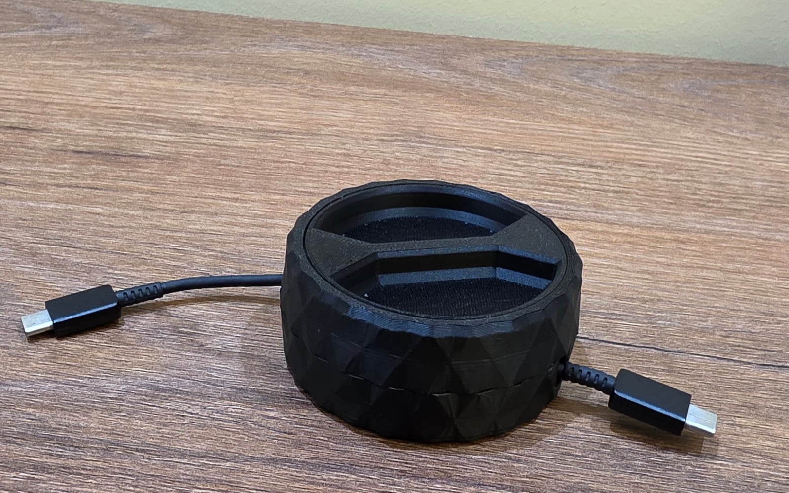 A black 3D-printed faceted round cable winder on a wooden desk, with a black charging cable fed through it.