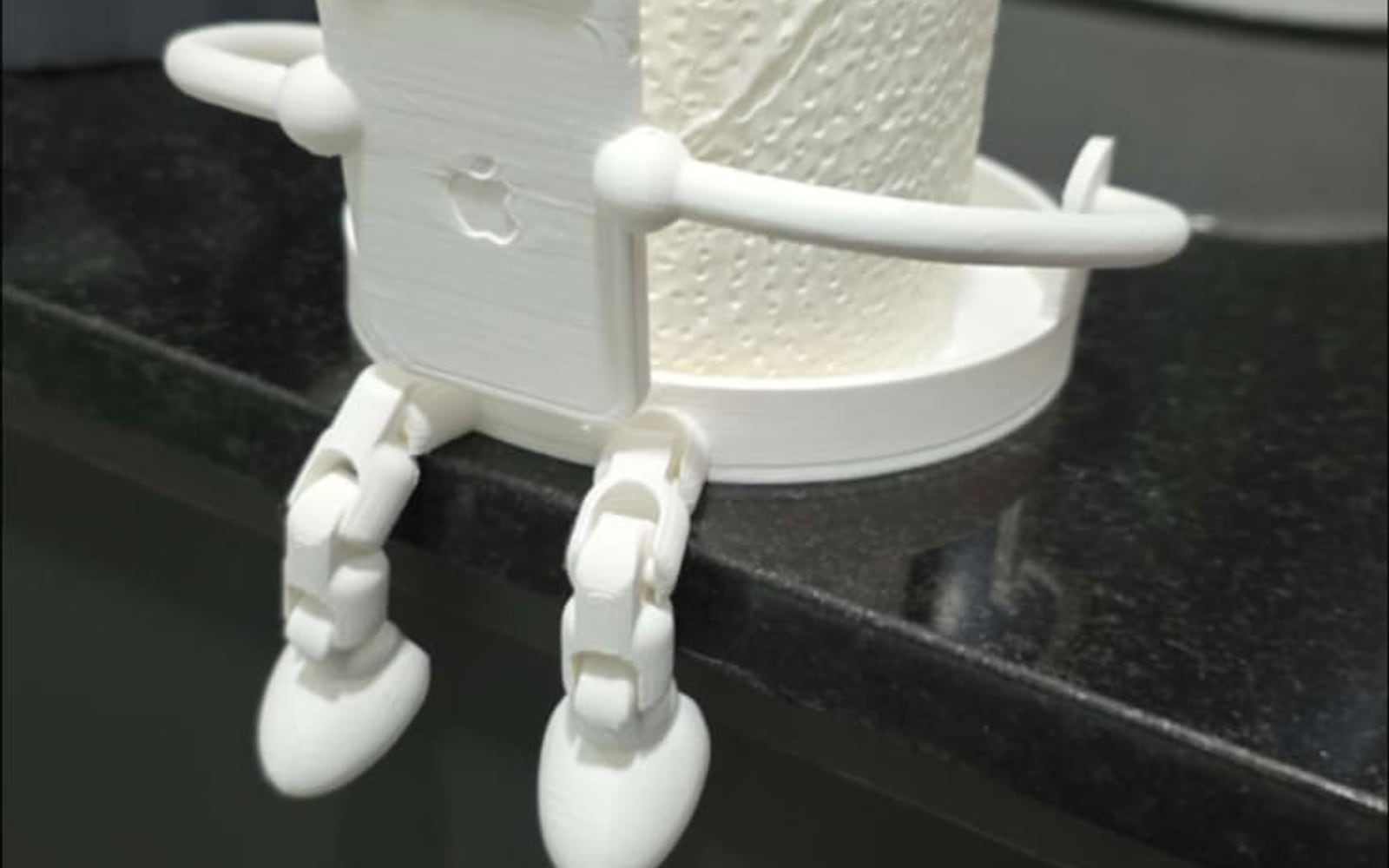 A white 3D-printed articulated robot figure sitting on a bathroom counter edge with its arms around a spare toilet roll, legs hanging over the side.